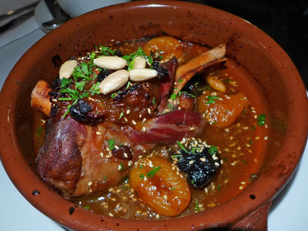 A red clay pot contains a lamb shank, dried fruit, and almonds.