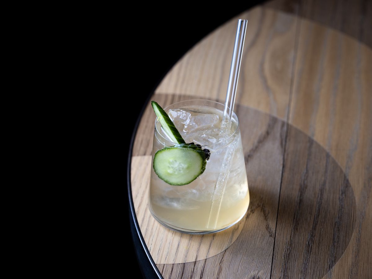 A cocktail garnished with cucumber, placed on a round wood table.