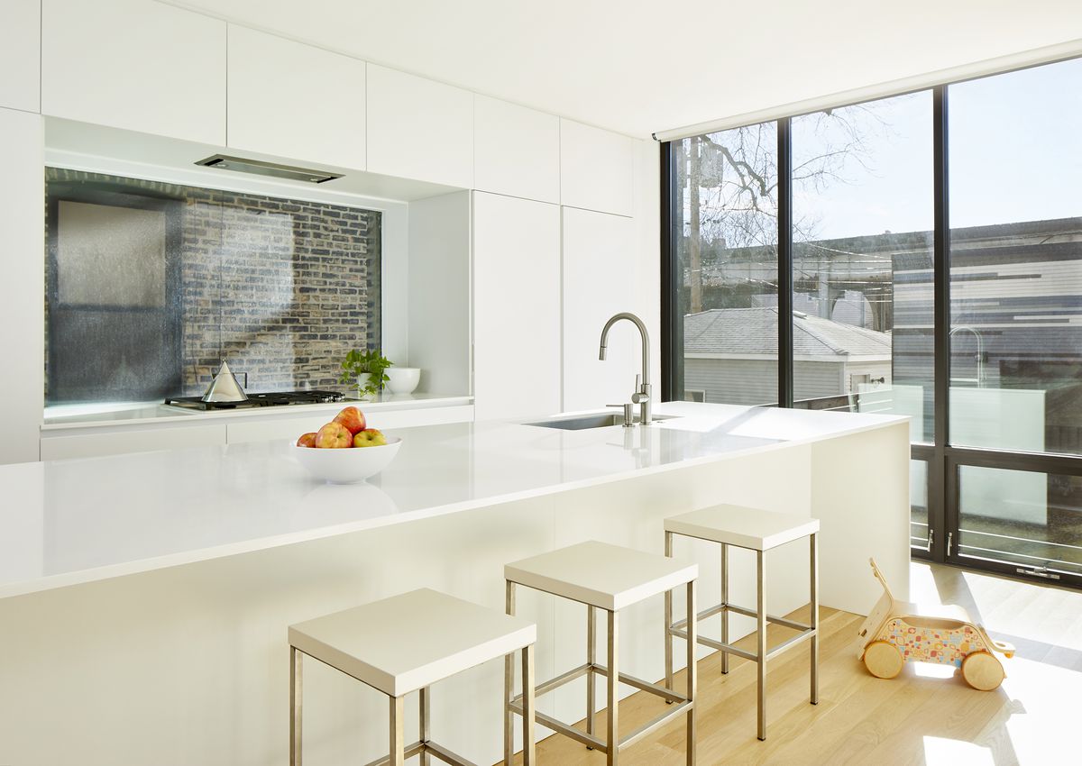 The kitchen is sleek and white, with no visible hardware or appliances. Through the windows, you see rustic brick and a garage with mulit-colored siding.