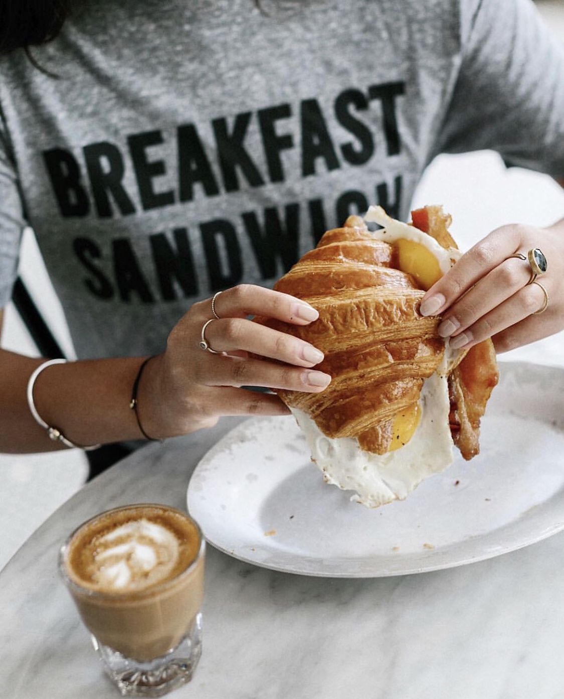 A woman wearing rings holds a croissant sandwich. Her shirt reads “Breakfast Sandwich.”