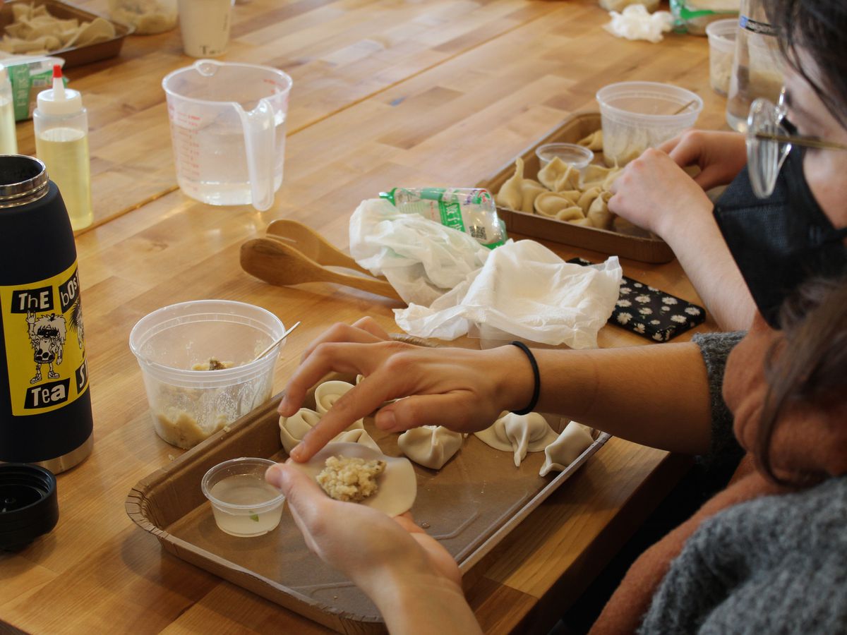 Hands are visible folding a dumpling dough sheet over a tray of dumplings with dough and fillings.