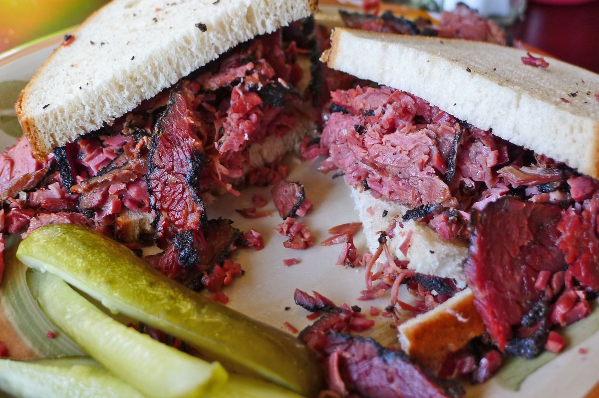 Two halves of a sandwich with pastrami tumbling out and pickles in the foreground.