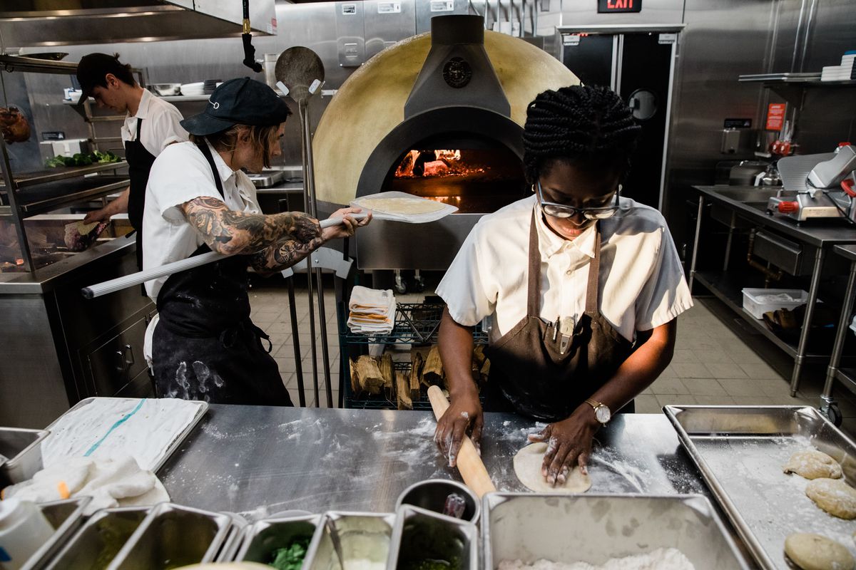 A female cook rolls out dough for flatbreads at a metal counter while a male employee uses puts a flatbread into a wood-burning oven.