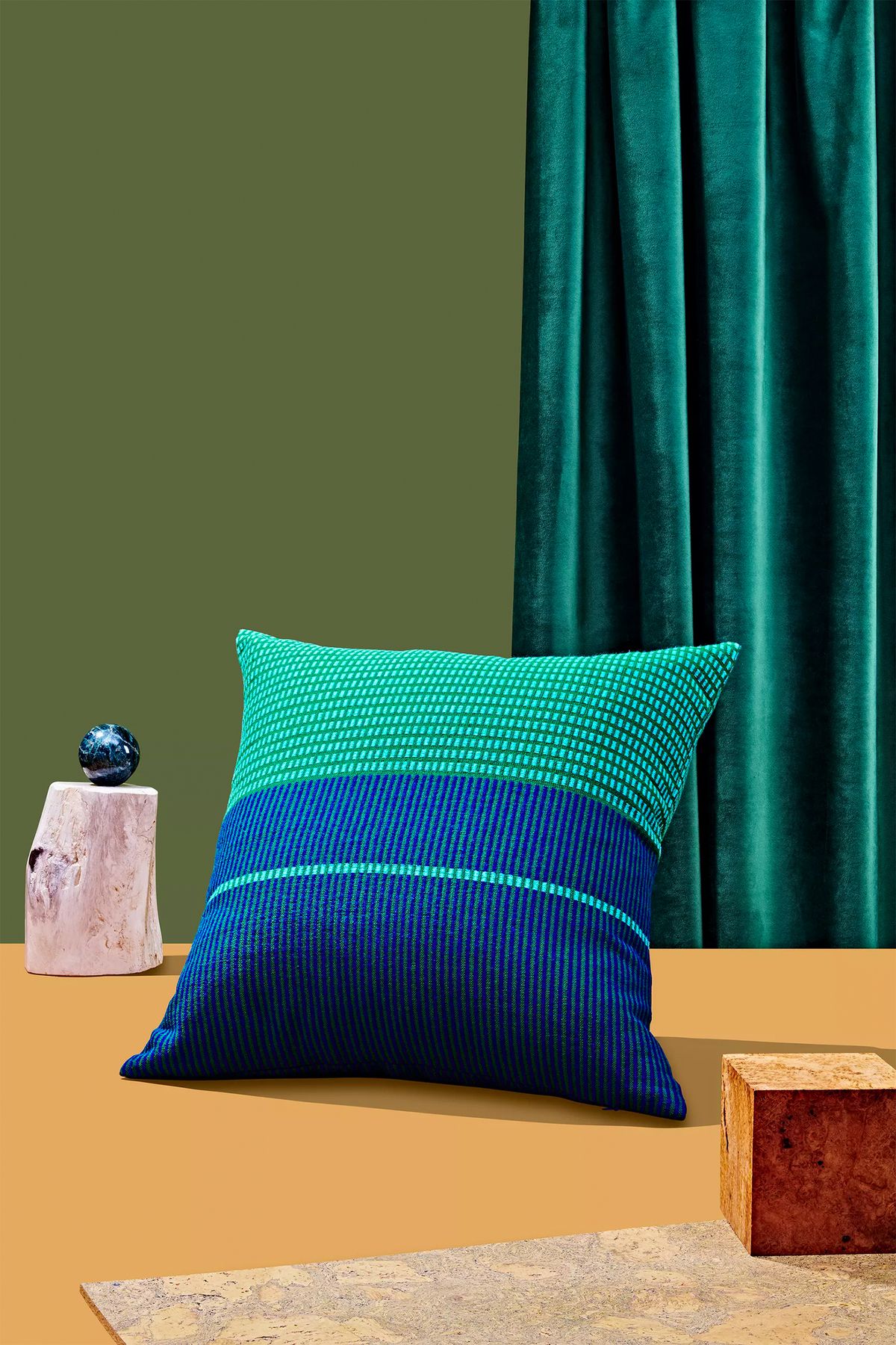 A green and blue handwoven pillow which is part of the Holiday Gift Guide 2019 for Curbed. The pillow is sitting upright on an orange surface. There are various design objects flanking the pillow. In the background is a green curtain and backdrop.