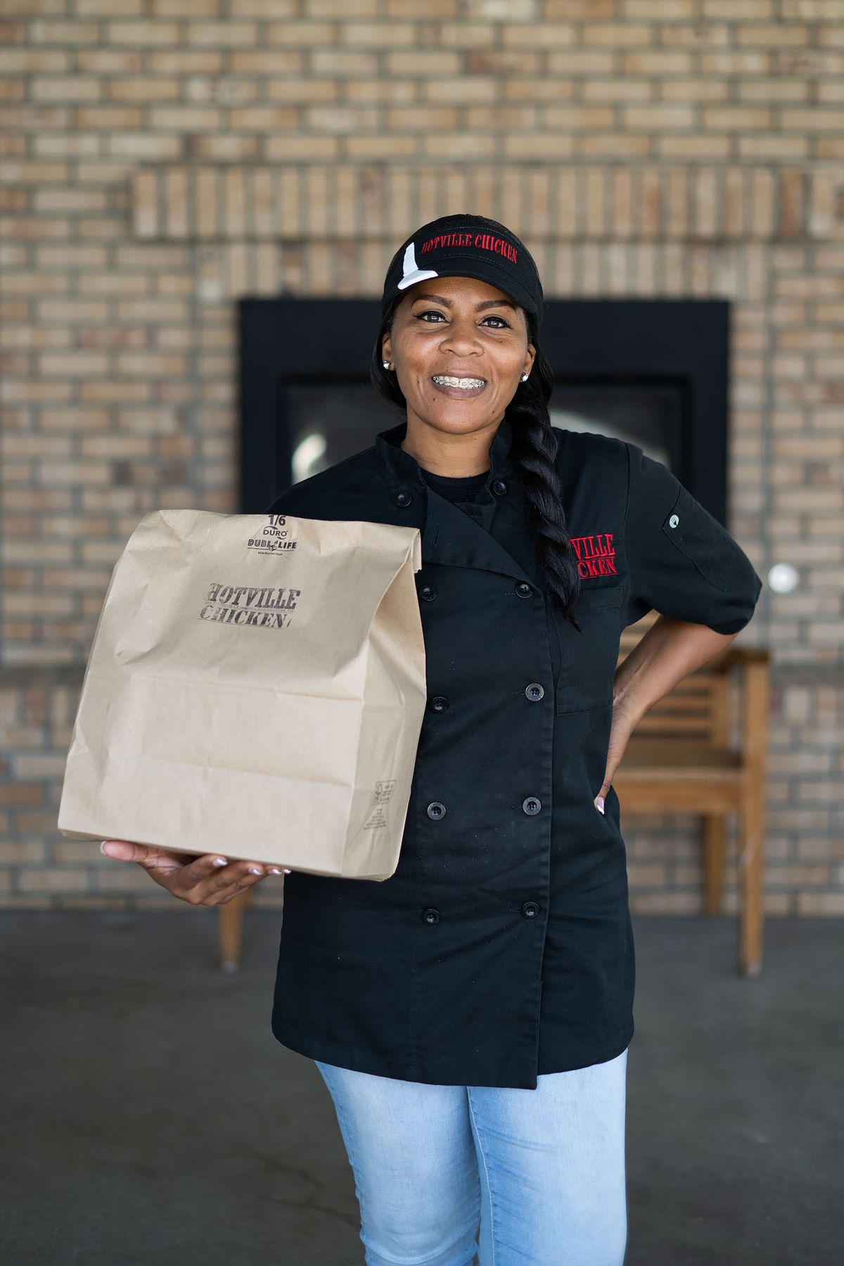 A Black woman wearing a black kitchen uniform holds a brown paper bag and smiles at the camera with a hand on her hip.