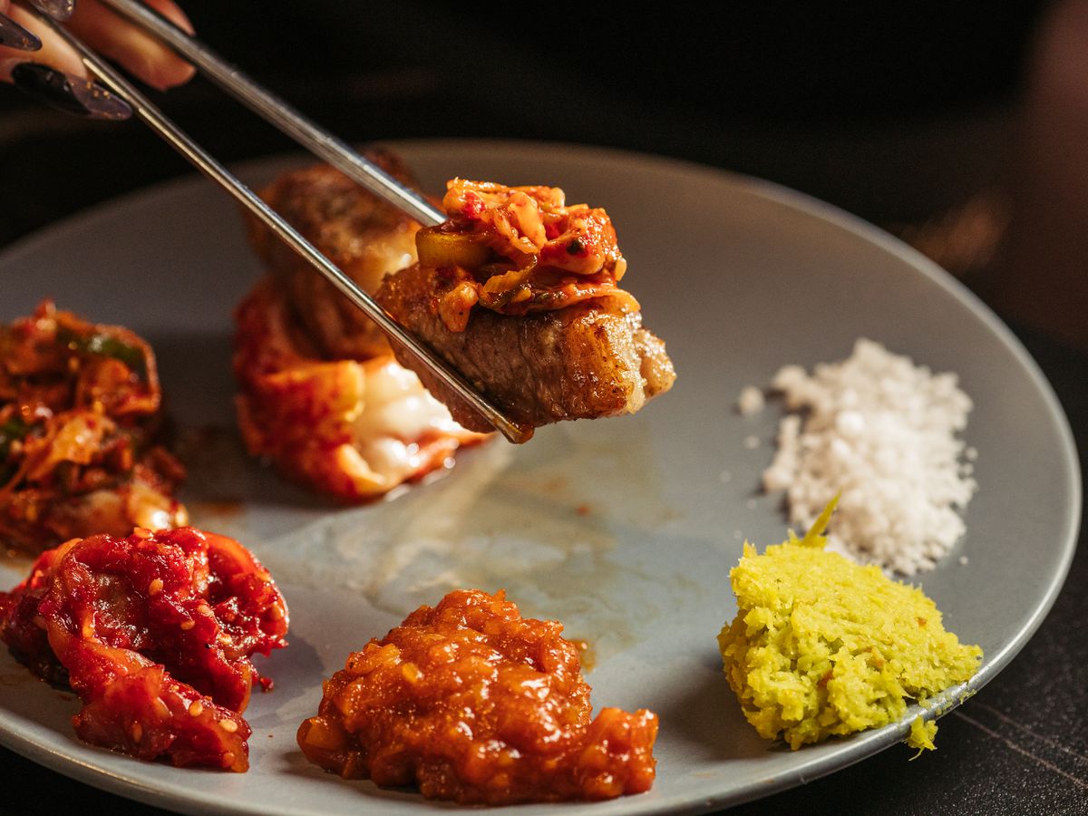Metal chopsticks hold a piece of grilled meat topped with kimchi in front of a plate of banchan.