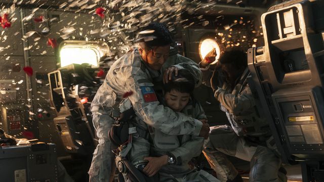 A bleeding man in an astronaut suit tries to cover the head of a woman in a similar suit as a series of windows in a small mechanical space shatter, spraying them and a third man with fragments of broken glass, in an action scene from The Wandering Earth II