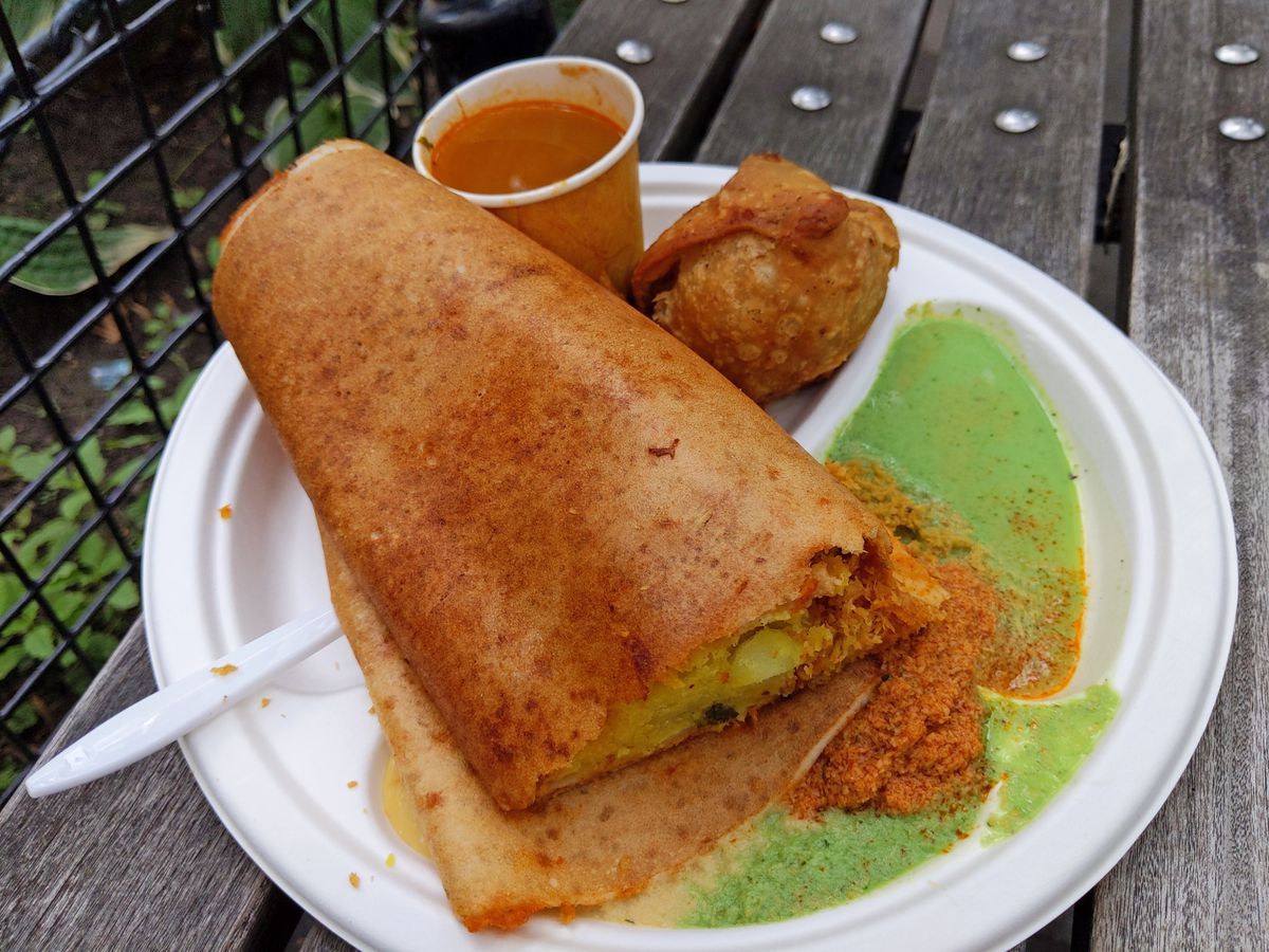 A white paper plate placed on a wooden bench with a dosa on it, a green cilantro sauce, a samosa, and a red sauce in a plastic cup.