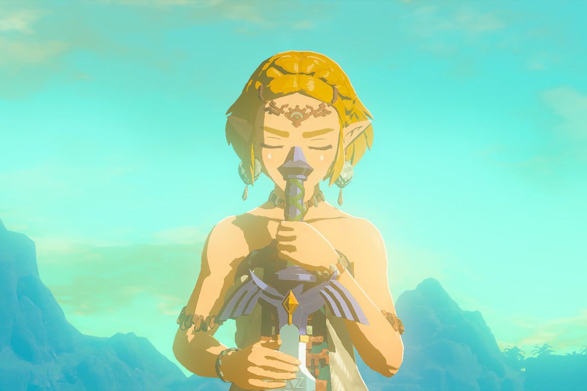 Zelda holds the Master Sword in Zelda: Tears of the Kingdom. She has her eyes closed, appears to be wearing ceremonial clothing, and we can see sky and mountains behind her. 