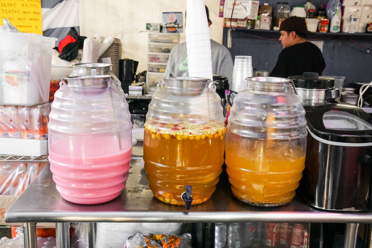 Three clear jugs hold different drinking juices, including a bright pink option, atop a metal table at a daytime street food vendor market.