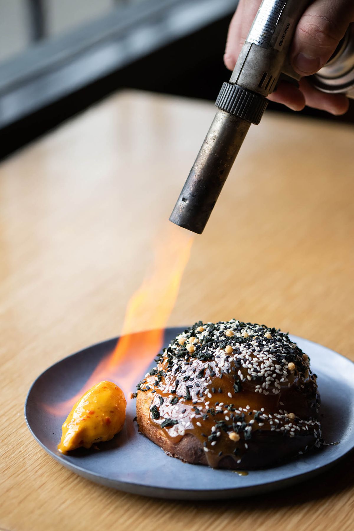 A blowtorch heats up butter next to a seeded roll on a plate.