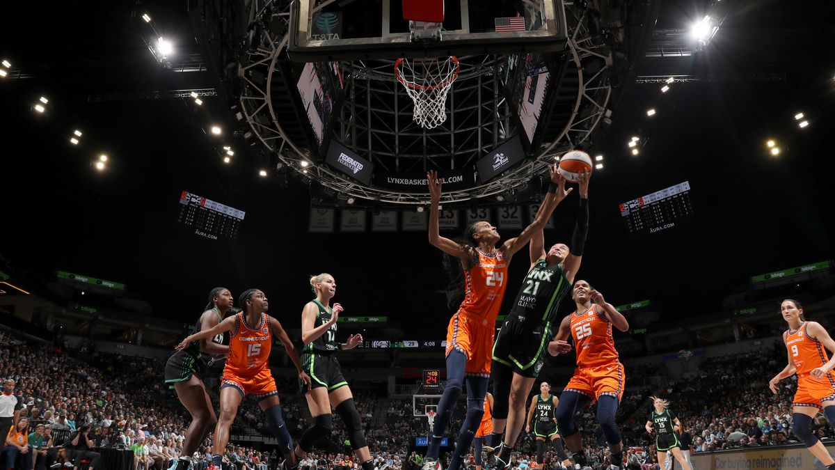 Women in orange uniforms and women in black uniforms playing basketball in front of a large audience. The shot is from behind the hoop; one player jumps to rebound as another tries to block her. 