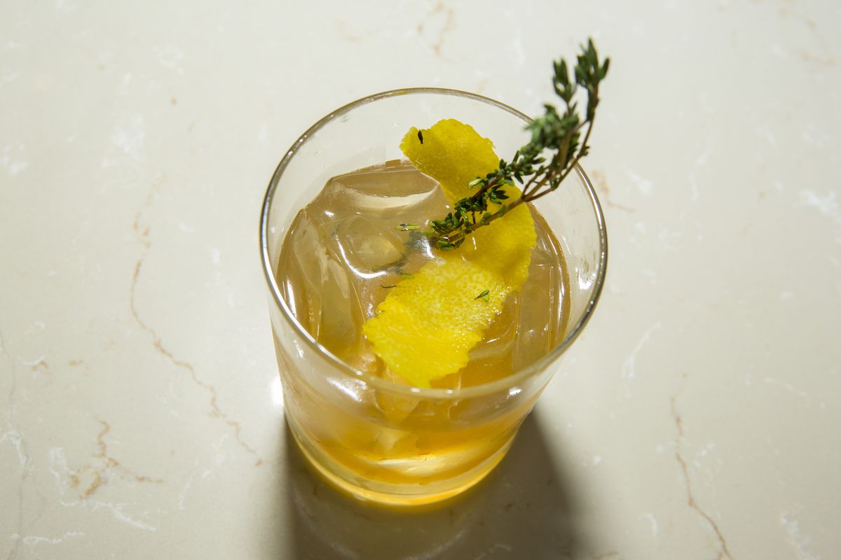 A rocks cocktail glass contains a yellow drink with ice, a twist of lemon peel, and a sprig of green thyme.