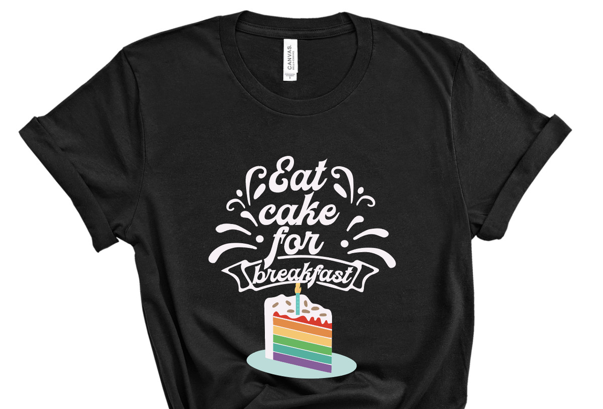 A black shirt that says eat cake for breakfast with an illustration of a slice of rainbow cake with a birthday candle.