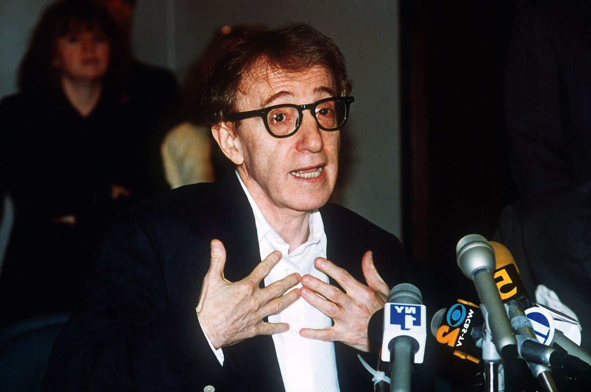 Woody Allen stands before a podium covered in microphones, speaking and gesturing to himself.