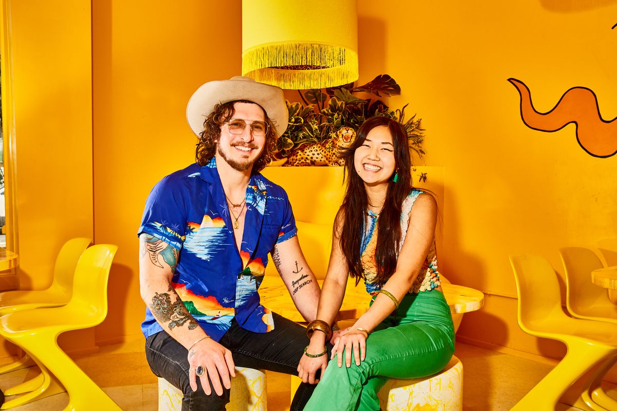 A couple is sitting in a bright yellow room.  He wears sunglasses, a cowboy hat and a button-up shirt patterned like a sky with rainbows.  She wears a tie-dye top and green pants.