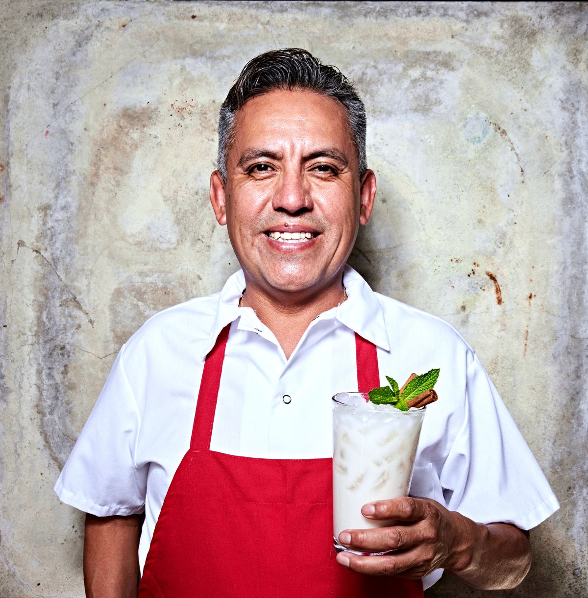A man in a white shirt and red apron holding up a drink.