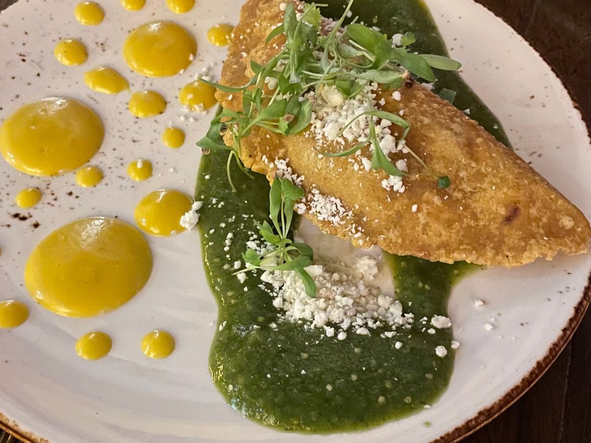 An empanada on top of a deep green salsa. Yellow dots of smooth sauce on the side of it and fresh crumbles of white cheese and microgreens on top of it.