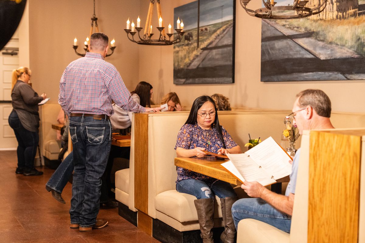 People sit, looking at menus and talking, in large u-shaped booths at a restaurant. Servers are at their tables. Chandeliers hang over them and large paintings of farms are on the walls.