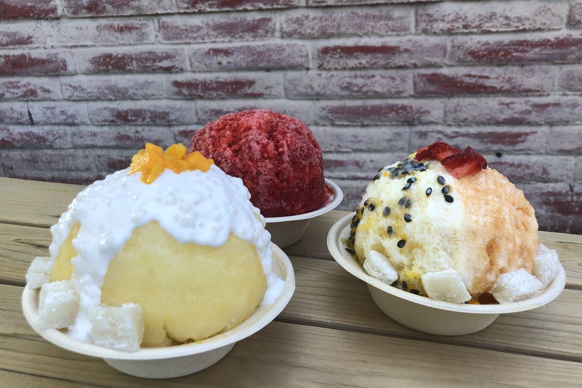 Three bowls of shave ice on a light wood table in front of a red brick wall. 