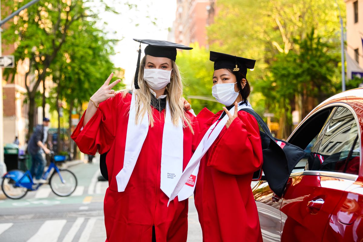 Two women flash peace signs as they pose together in red caps and gowns, their faces covered with masks. 