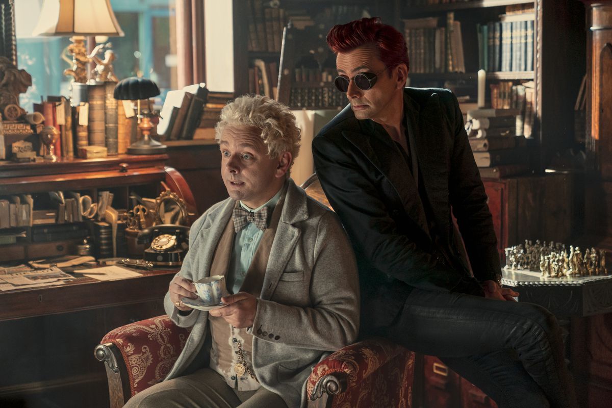 Aziraphale (Michael Sheen) sits in a chair with Crowley (David Tennant) leaning over his shoulder; they are both looking surprised at something