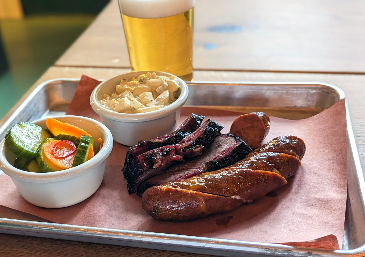 Sausages and slices of brisket on a tray beside cups of sides and a glass of beer.
