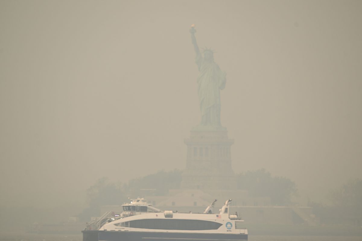 A ferry is seen in relatively clear view passing in front of the Statue of Liberty in New York City, which is obscured in a red-brown haze of smoke.
