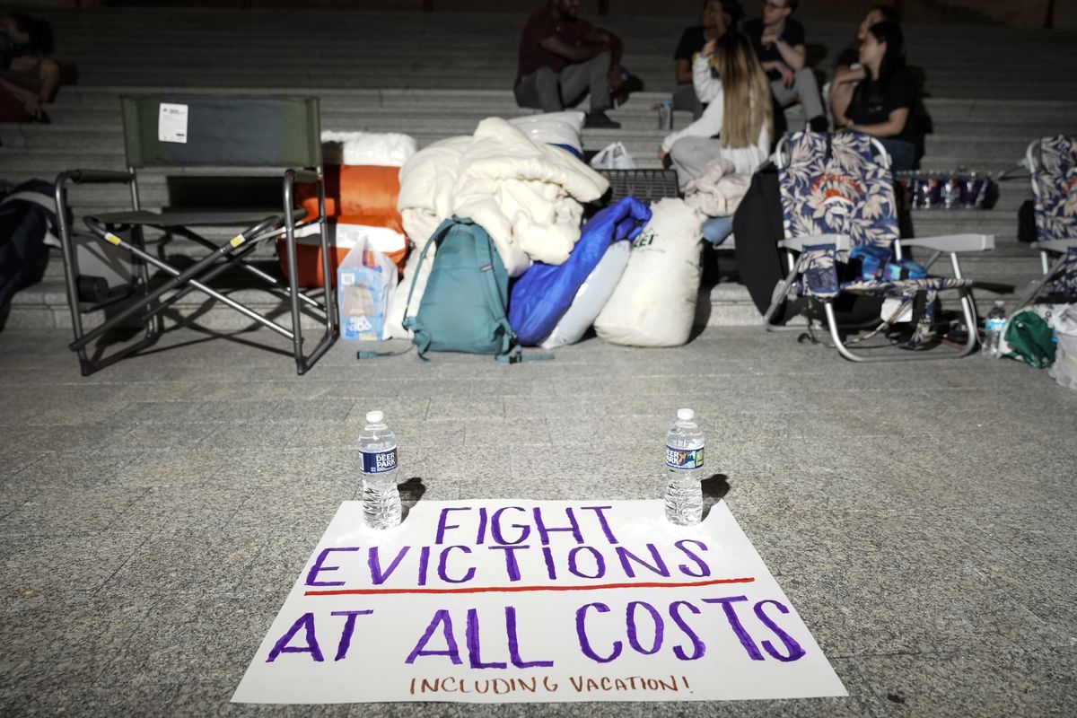 A sign calling for fighting evictions is set on the ground in front of outdoors chairs, bags and water bottles, where people have been camping outside.