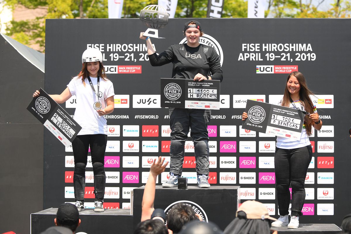 Hannah Roberts stands atop a podium at the 2019 UCI BMX Freestyle World Cup, holding the competition’s trophy and a fake check for her prize winnings. The second- and third-place finishers are on the podium with her.