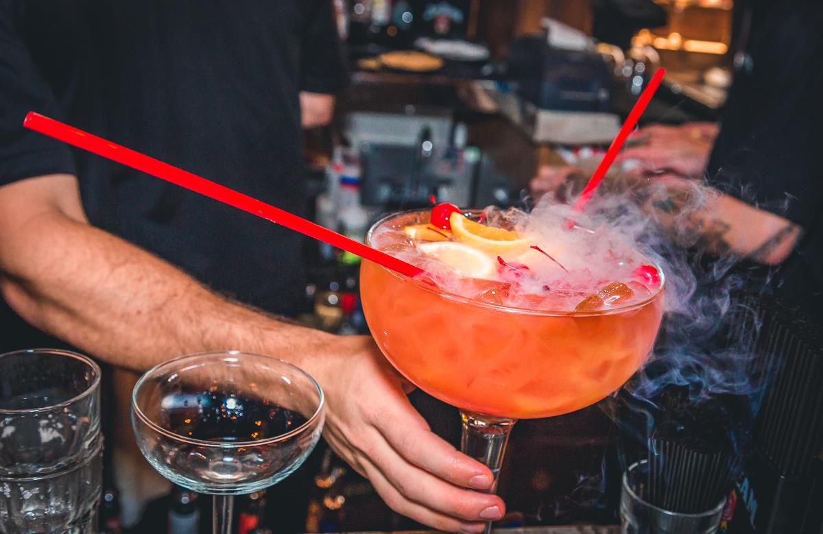 A bartender holds a pink scorpion bowl in a giant glass. It has two straws coming out of it.