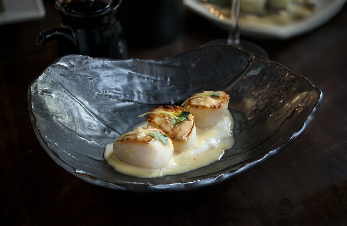 Three seared scallops sit in a creamy pool of pale yellow sauce in a decorative black bowl.