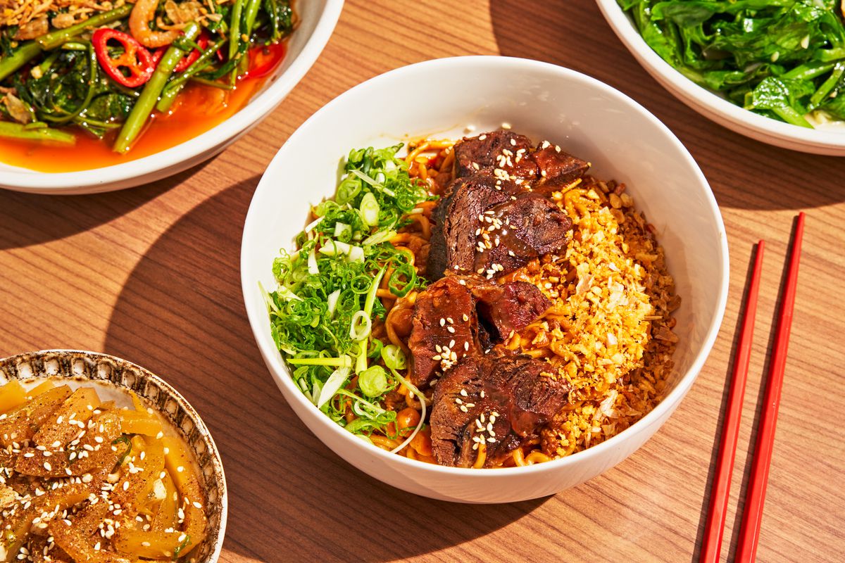 A photo features a white bowl filled with noodles and beef, next to a small bowl with celtuce salad and red chopsticks beside it.
