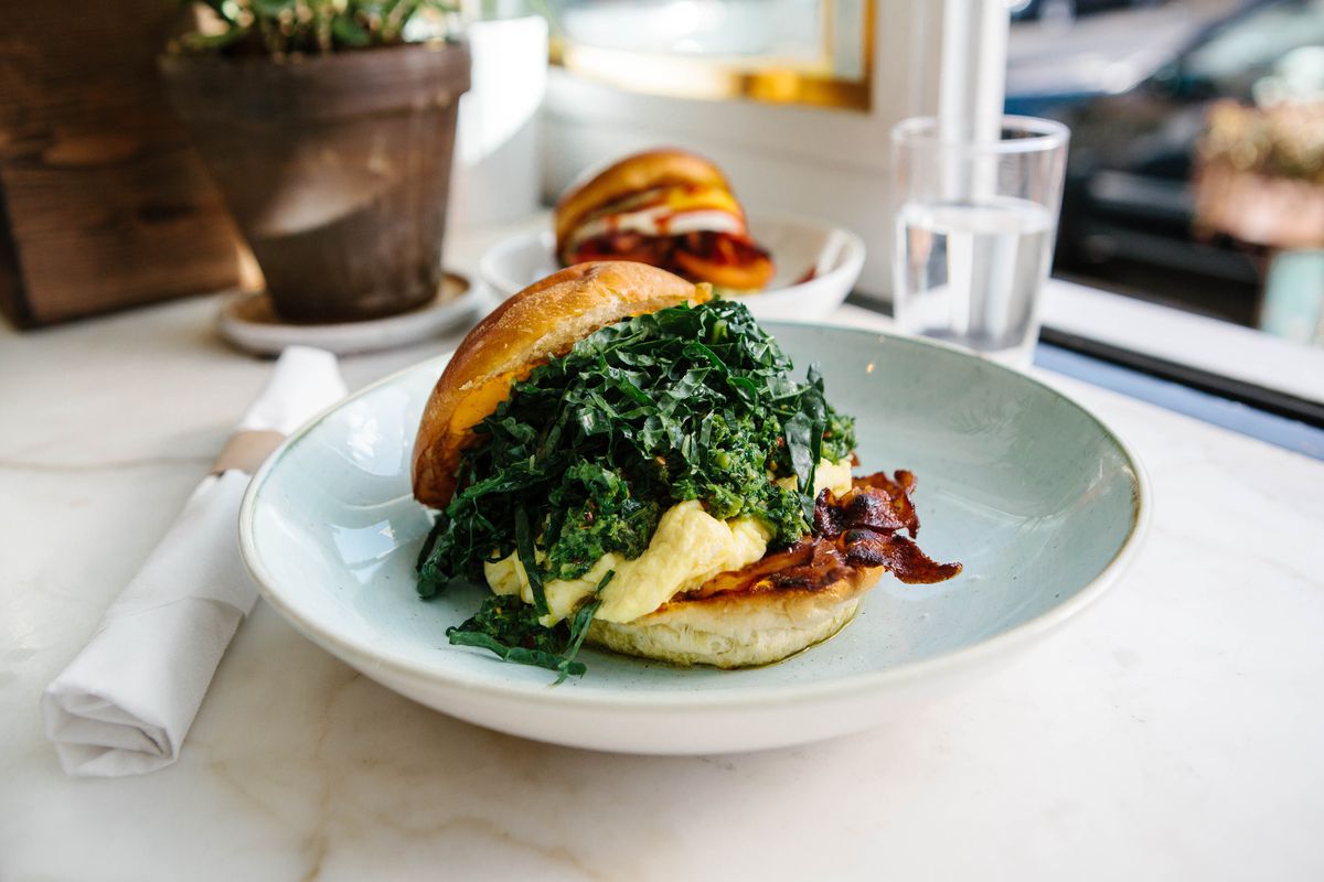 In the foreground, a sandwich topped with bacon, egg, and a mountain of kale rests on a toasted bun. In the background, tableware and a similar sandwich are visible.