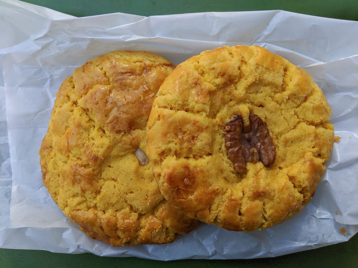 Two large cookies —&nbsp;one with an almond in the center and one with a walnut —&nbsp;overlap on a white paper bag on a green table