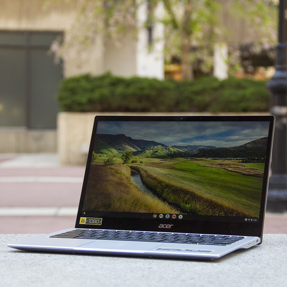 The Acer Chromebook Spin 513 sits at an angle to the left, open on a stone bench.  The screen displays a rustic view.