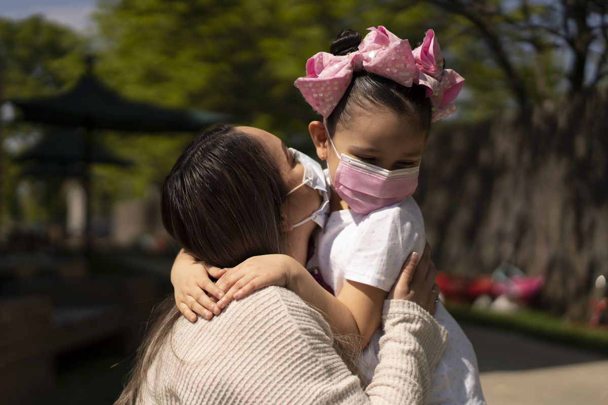 A mother wearing a tan shirt and a protective mask holds her daughter, who is wearing a white shirt, pink mask and pink polka-dot bow. The background is a swirl of green trees.