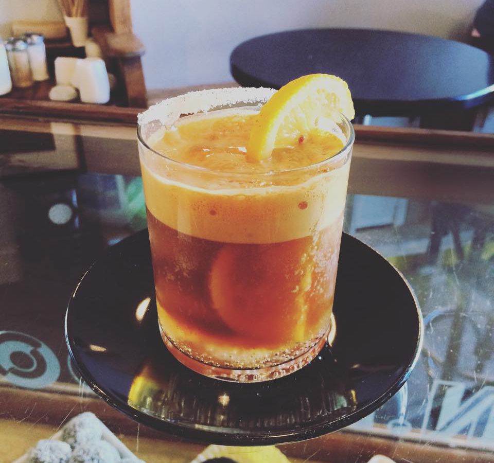 A frothy glass of espresso and tonic with a thick head and an orange wheel perched on the rim sits on a glossy saucer on a glass counter