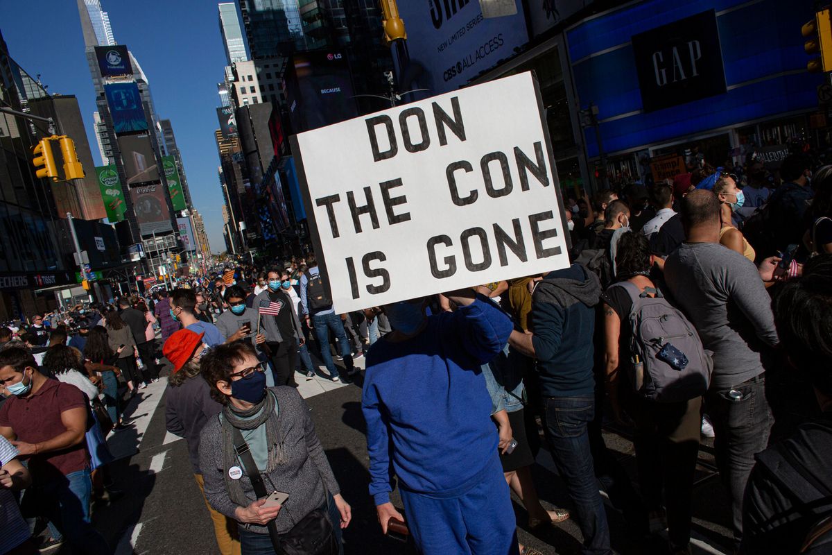 With Times Square glowing in the background, a protester holds a sign reading “Don the Con is Gone.”
