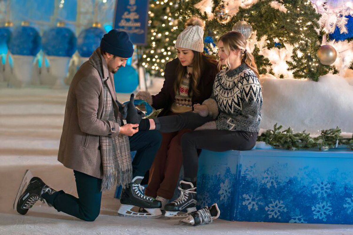 A man kneels to help two seated women put on ice skates in a winter setting.