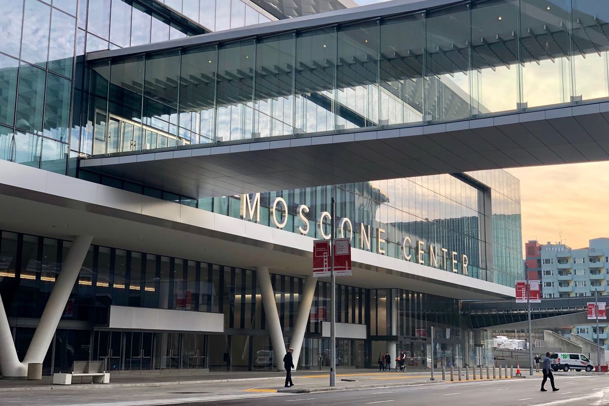 A glass-enclosed pedestrian bridge leading into the second story of a broad building with lettering out front reading “Moscone Center.”