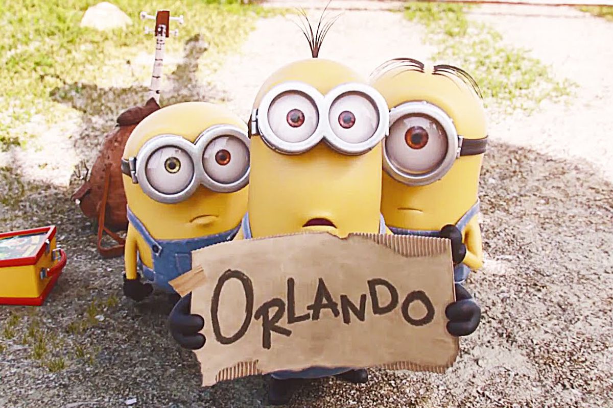 Three Minions holding a cardboard sign that says “Orlando,” as they hitchike