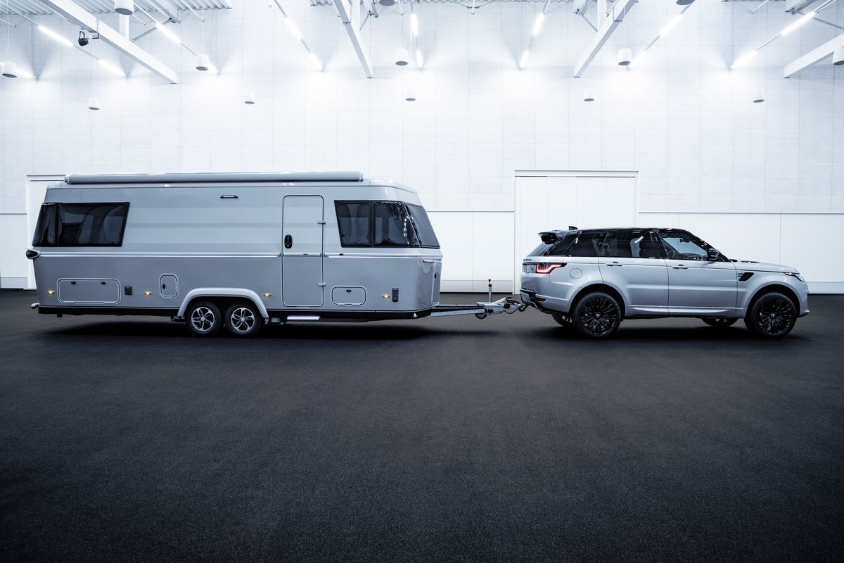A blue camper trailer is attached to a blue van. The trailer and van are in a large white showroom.