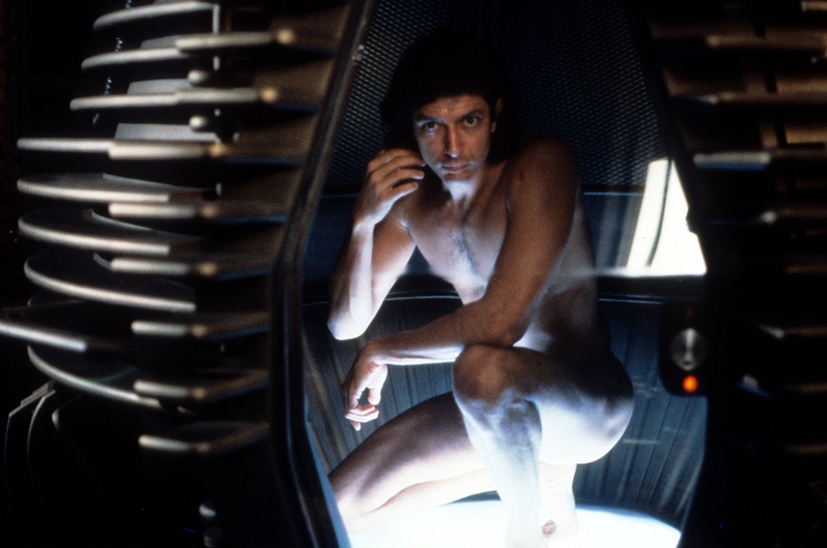 Jeff Goldblum, naked and harshly lit, crouches in a ominous, futuristic looking metal pod in a scene from the 1986 film The Fly