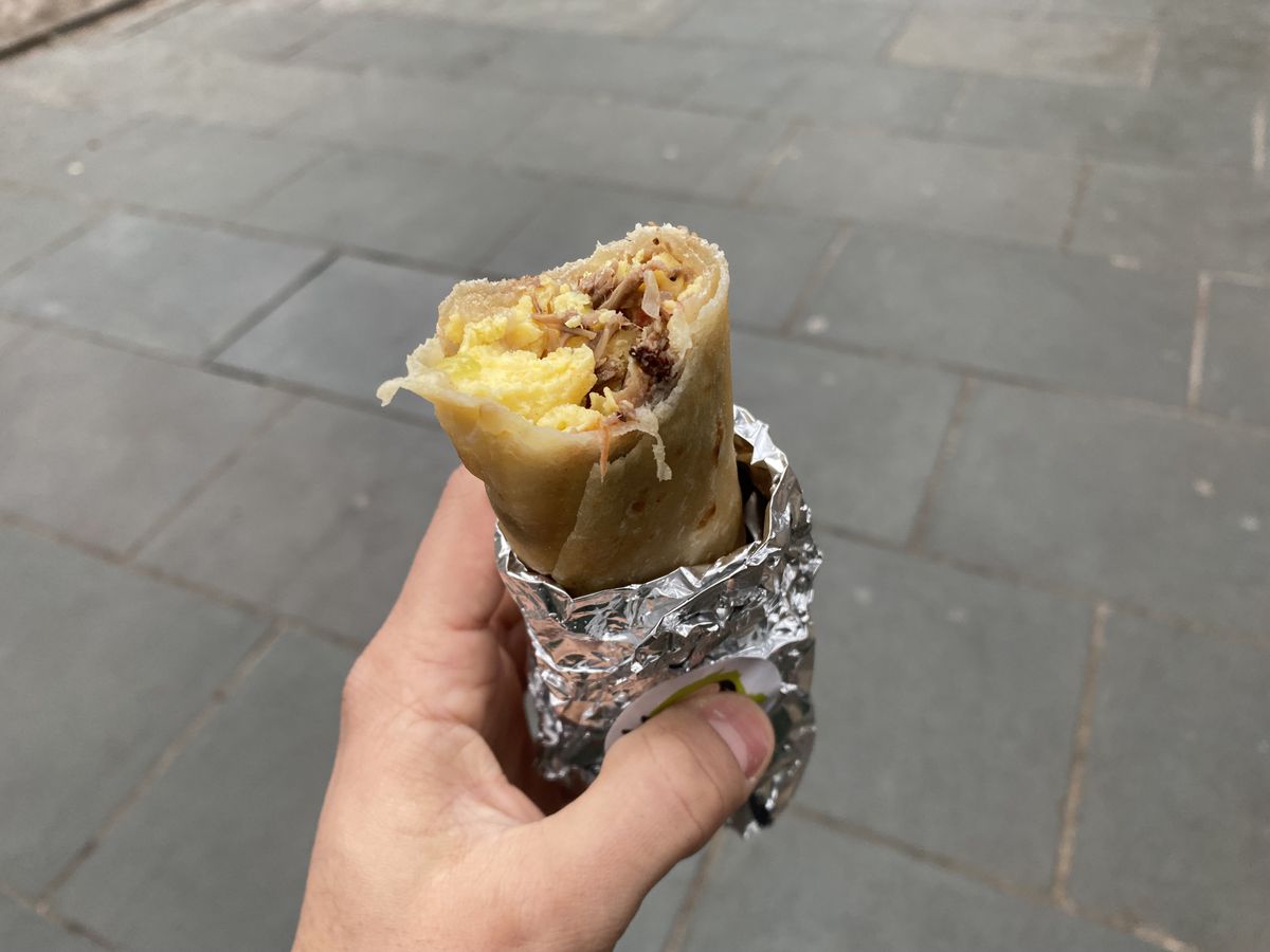 A hand holds a thin breakfast burrito, overflowing with pieces of egg and shredded machaca.