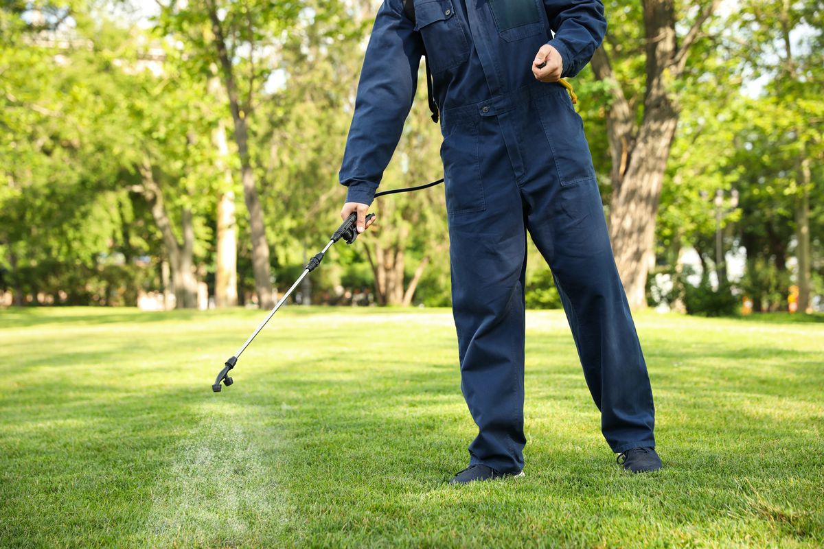 A pest control specialist wearing a navy blue jumpsuit uses a black wand to spray pest control solution on a bright green yard. 