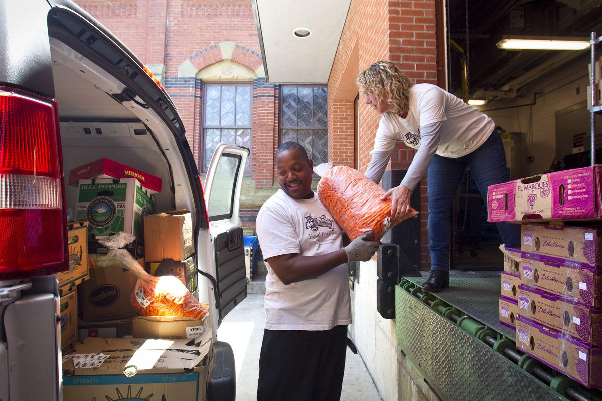 A woman standing at a loading dock passes a bag of food to a man below; he is about to load it into the back of a van full of boxes and bags of food