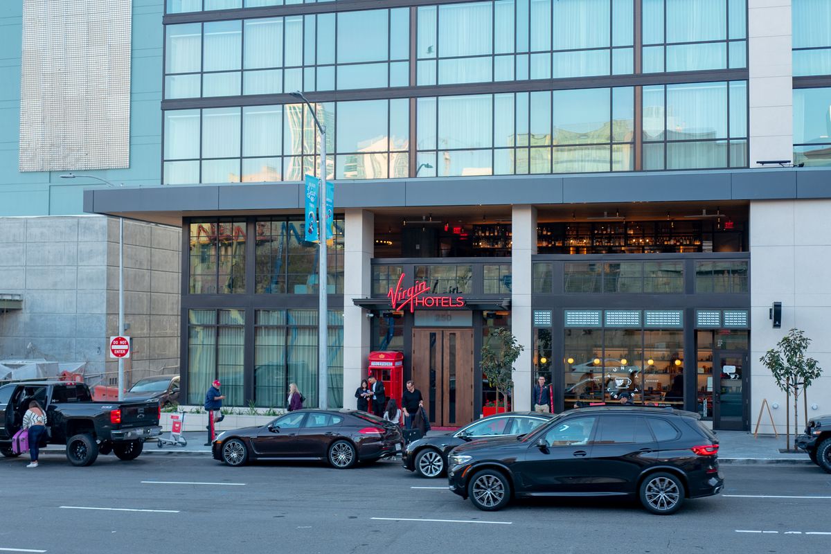 People walking on a sidewalk in front of a building with banks of windows and a sign with a red logo that reads “Virgin.”