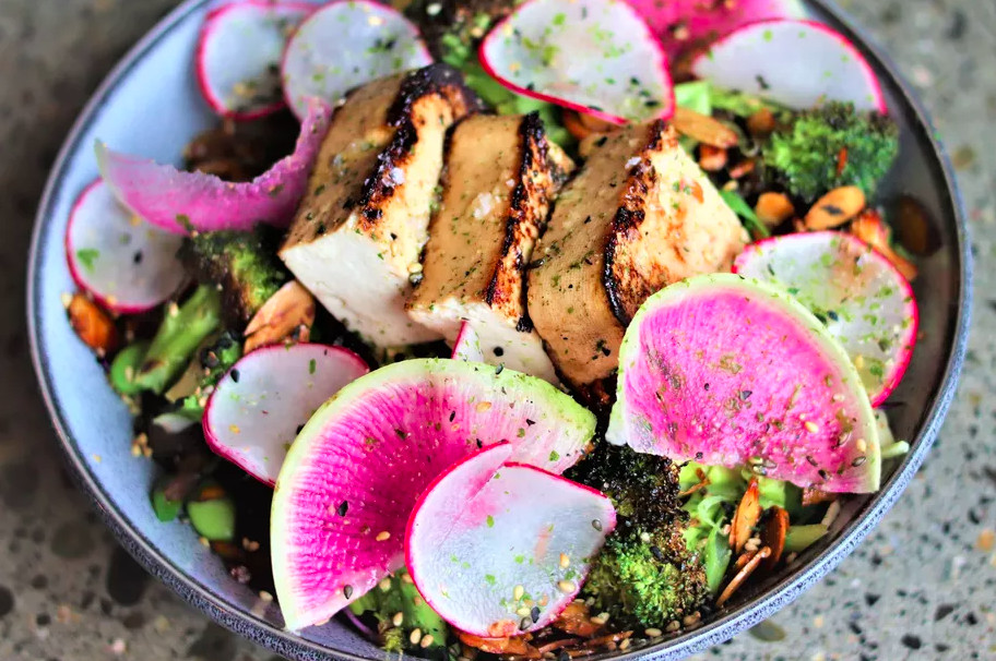A bowl of charred vegetables, radishes, and tofu sits on a table at Feel Good