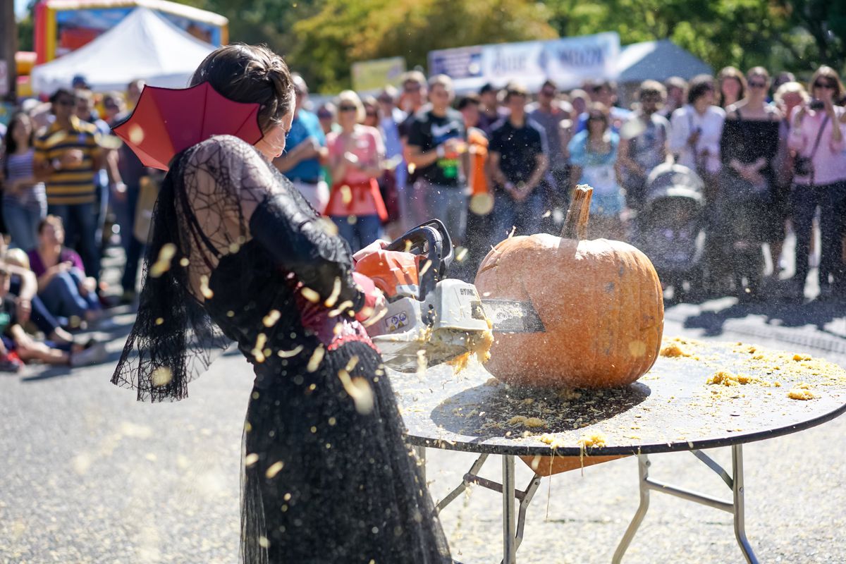 A woman dressed in goth clothes uses a chainsaw to carve a pumpkin as a crowd watches.